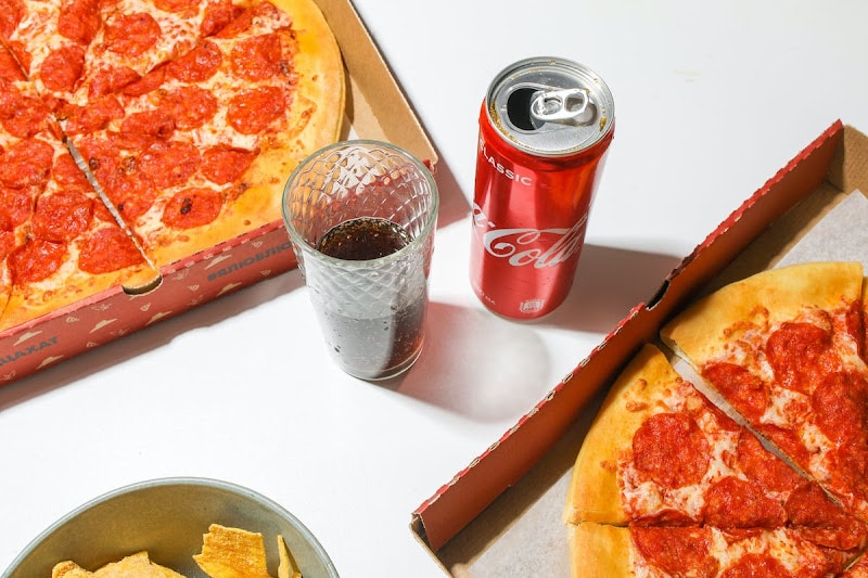 Coca cola can with a cup of soda in between two delivered pepperoni pizzas and corn chips.